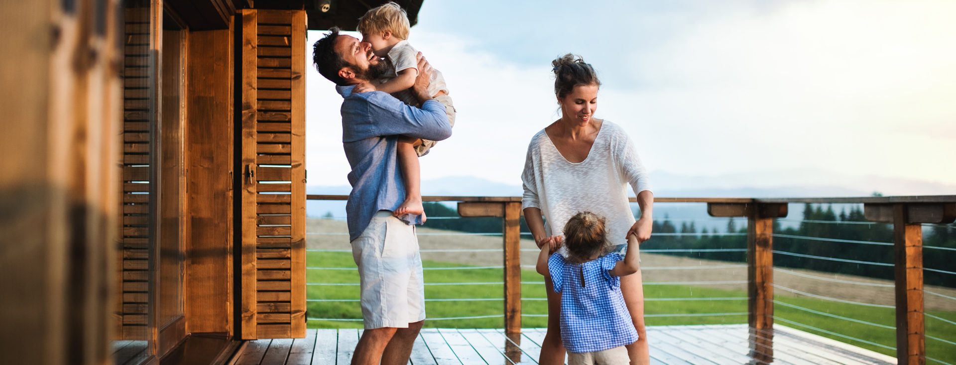 Family with happy small children playing in rain on patio by wooden cabin, holiday in nature concept.